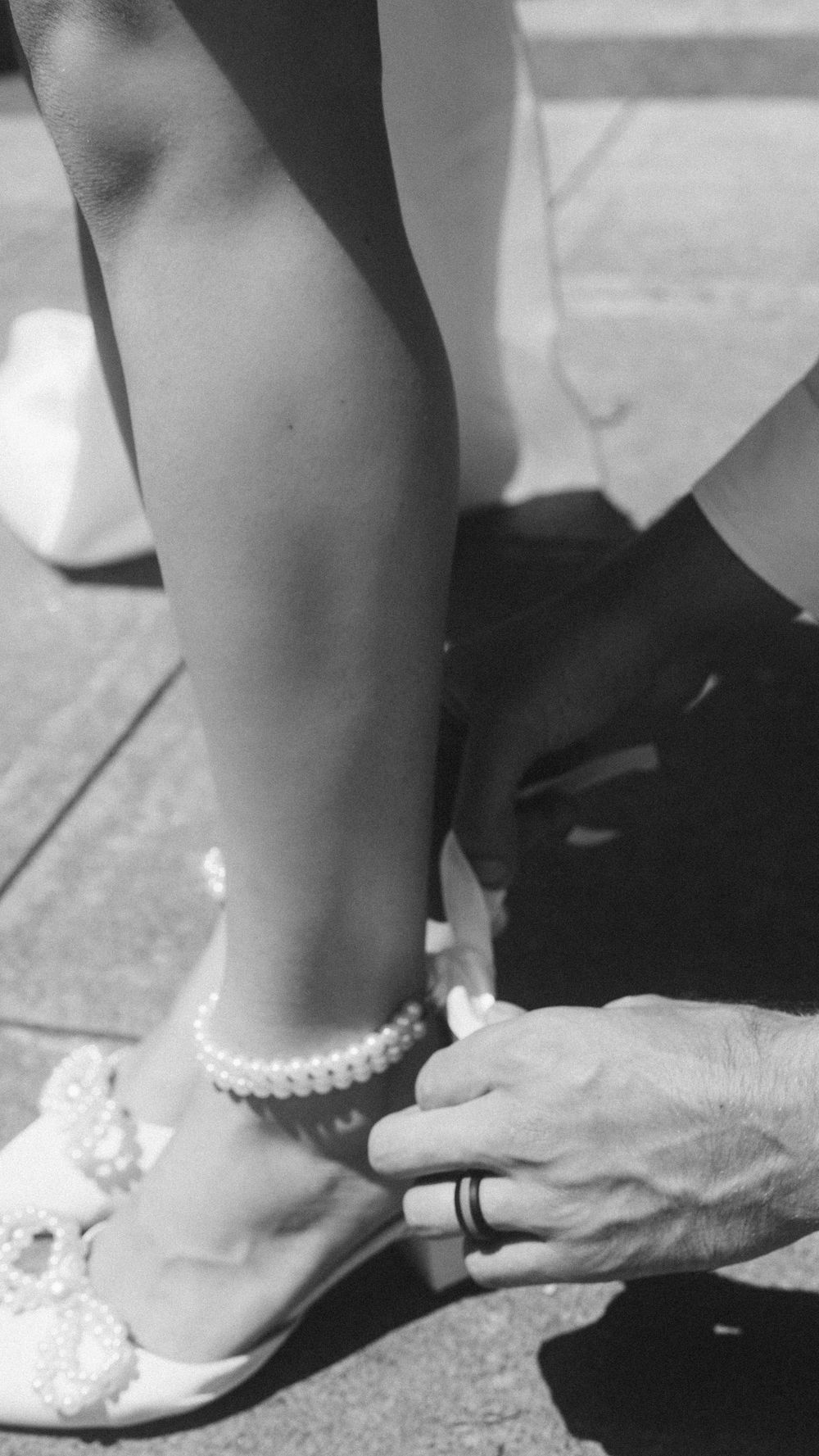 Black and white photo of a pearl anklet being fastened on a bride's ankle by an older hand wearing a ring.