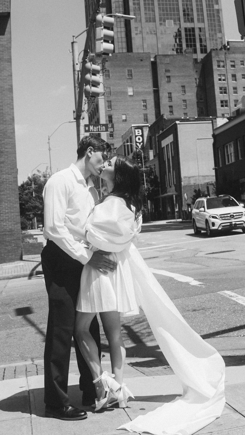 Black and white photo of a couple kissing in wedding attire on a city street corner with urban buildings in the background.