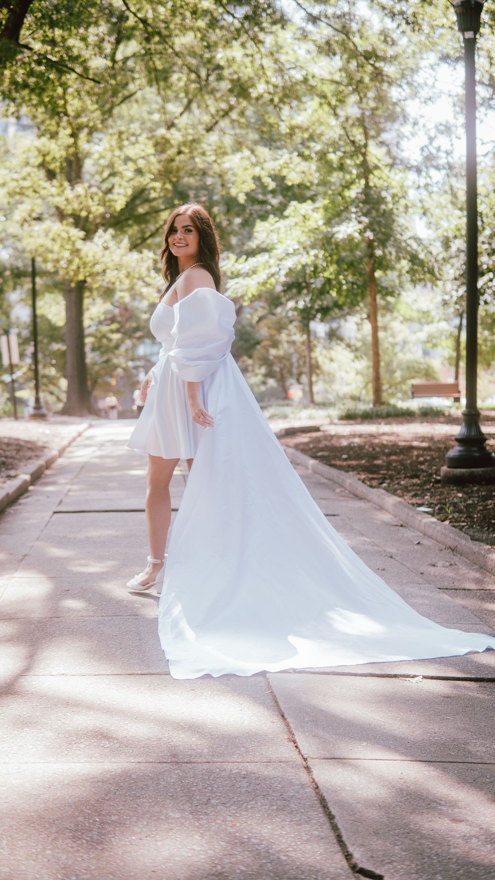 Smiling bride in a white off-shoulder mini dress with a long train poses on a sunlit park pathway.