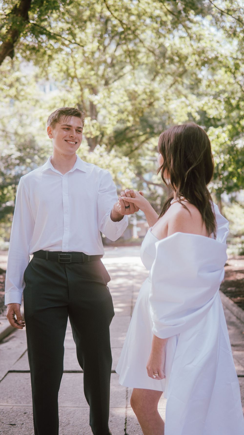 Smiling groom in white shirt and black pants holds hands with bride in white off-shoulder dress outdoors.