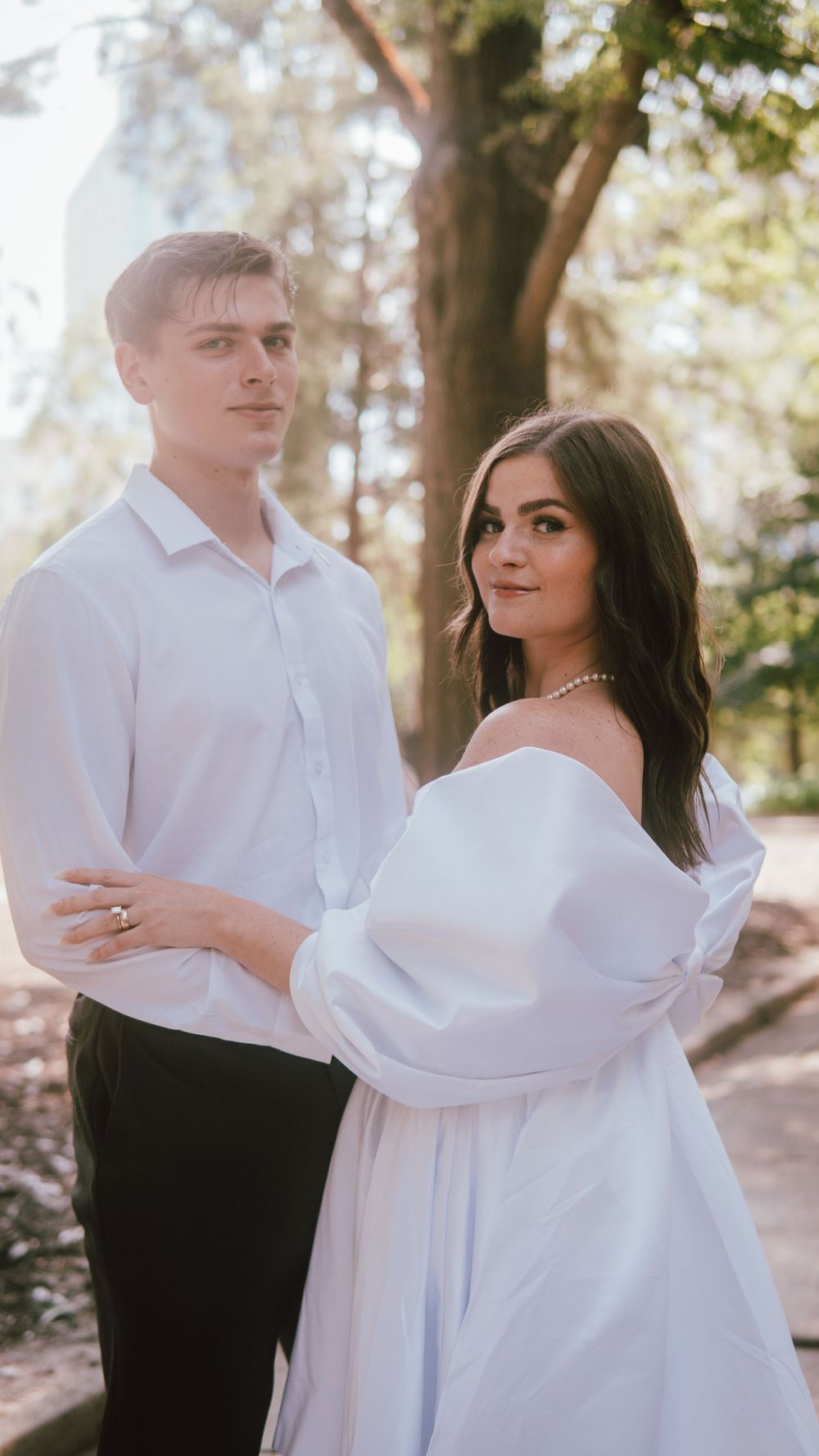 A couple in white outfits poses outdoors, woman in off-shoulder dress with pearl necklace, man in white shirt.