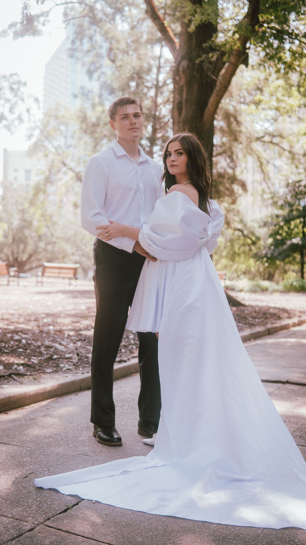 A couple in wedding attire poses in a sunlit park, the bride in a white off-shoulder gown with a long train.