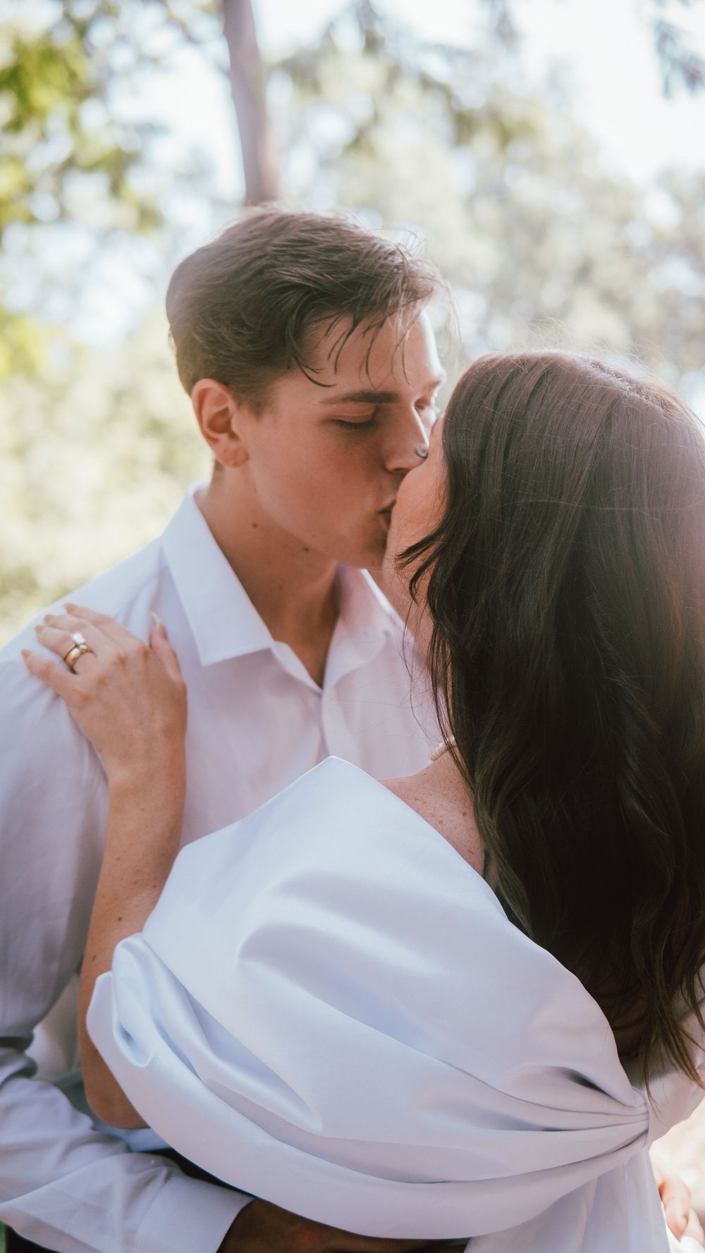 A couple sharing a romantic kiss outdoors, dressed in white, with lush green trees in the soft background.
