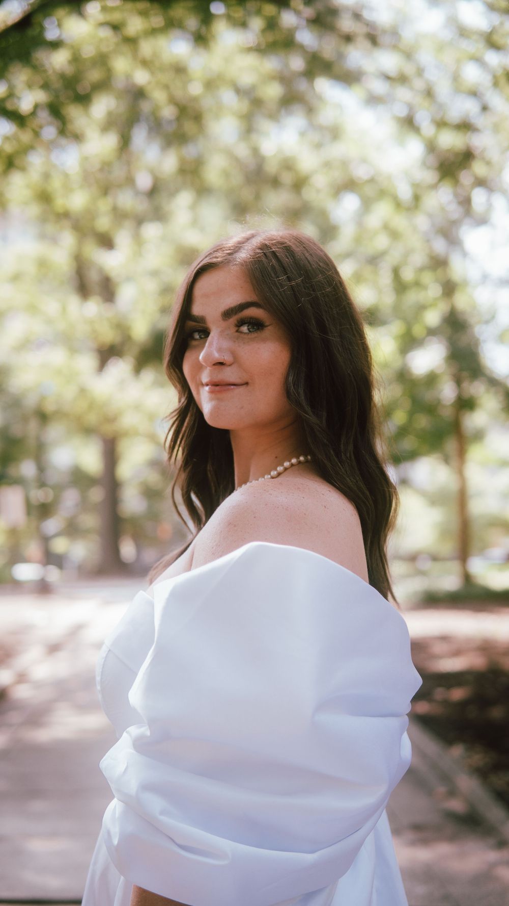 Young woman in white off-shoulder dress with pearl necklace posing outdoors among trees.