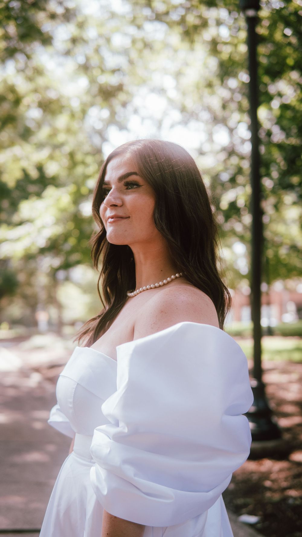 Young woman in white off-shoulder puff sleeve dress and pearl necklace posing outdoors in a sunlit park.