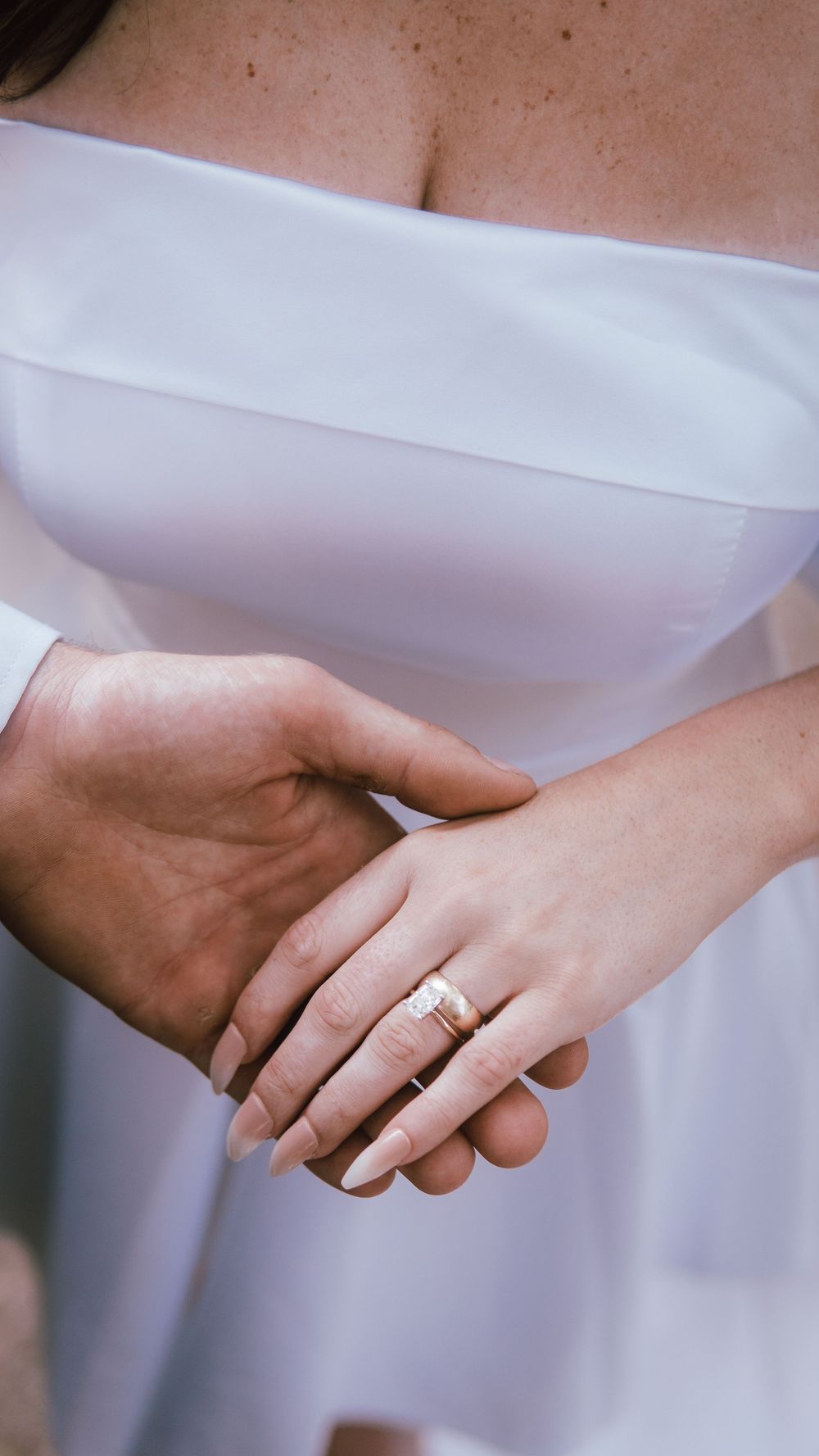 Close-up of bride and groom holding hands, showcasing wedding rings on a white off-shoulder bridal gown.