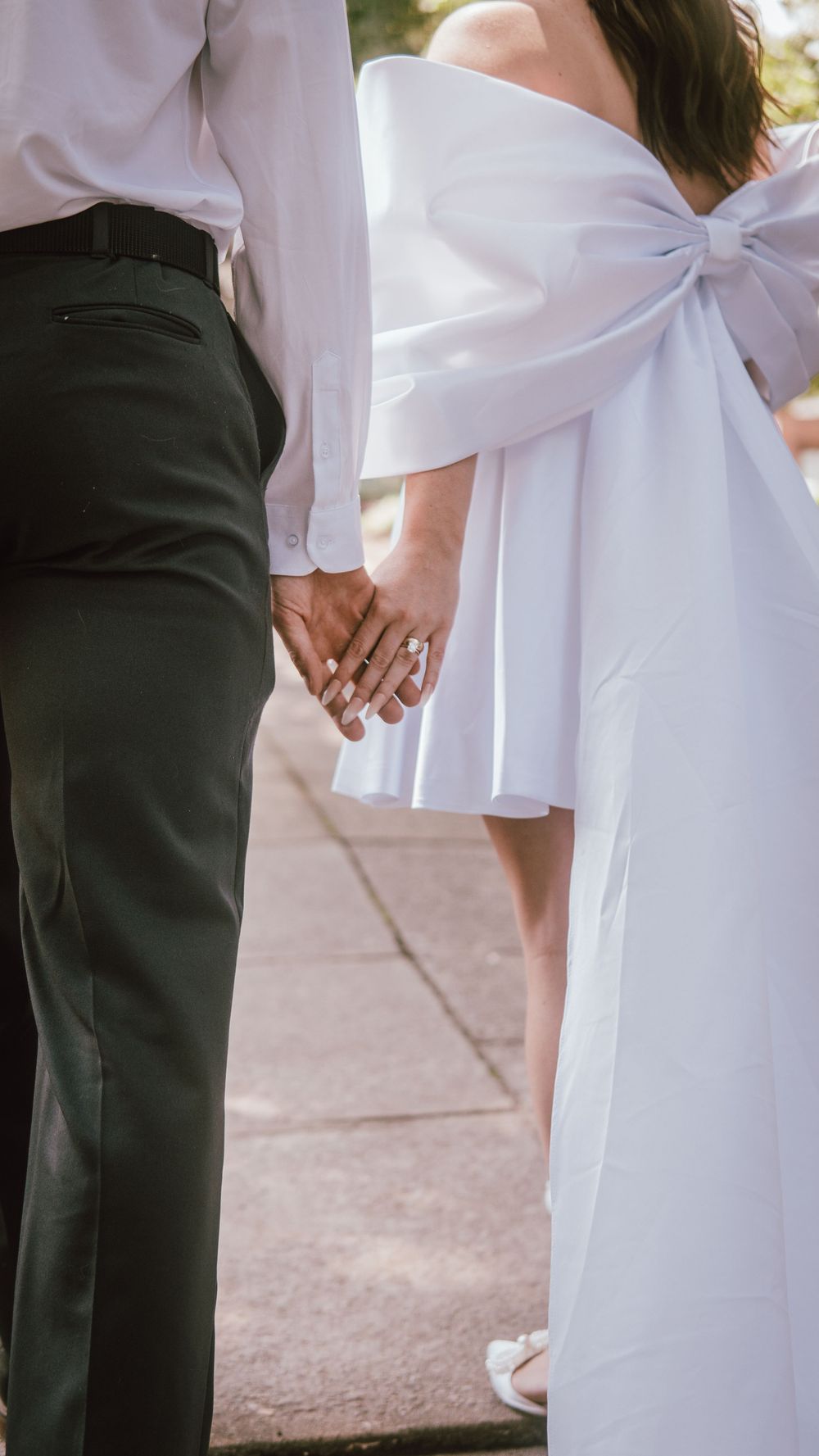 Bride in white off-shoulder gown with bow holds hands with groom in black trousers on wedding day.