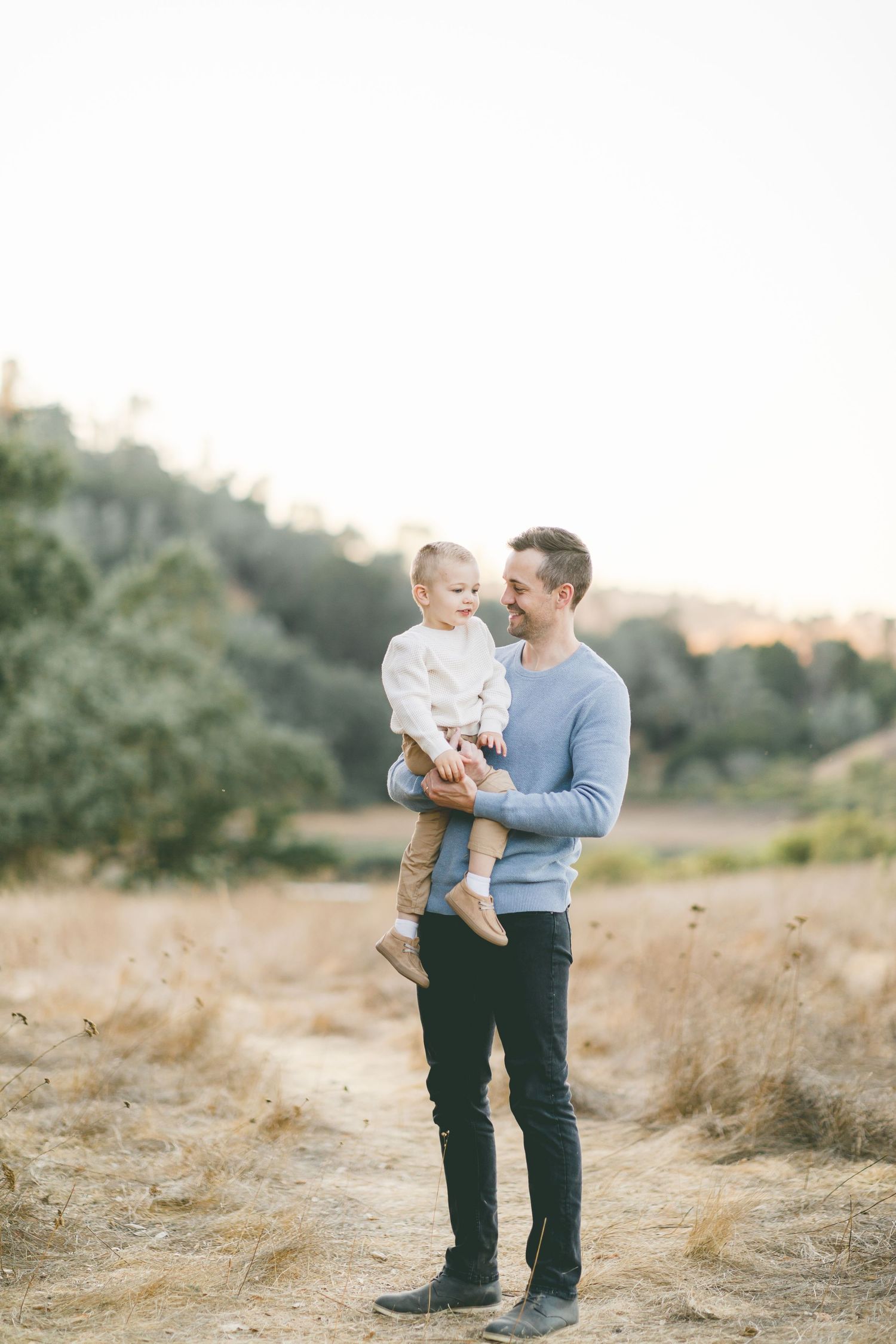 A smiling man in a blue sweater holds a toddler in a sunny, dry field with green hills in the background.
