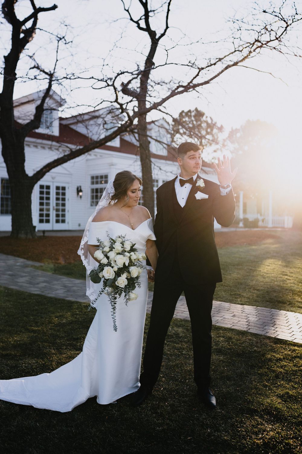 Bride in white off-shoulder gown with bouquet and groom in black tuxedo pose outdoors at golden hour.