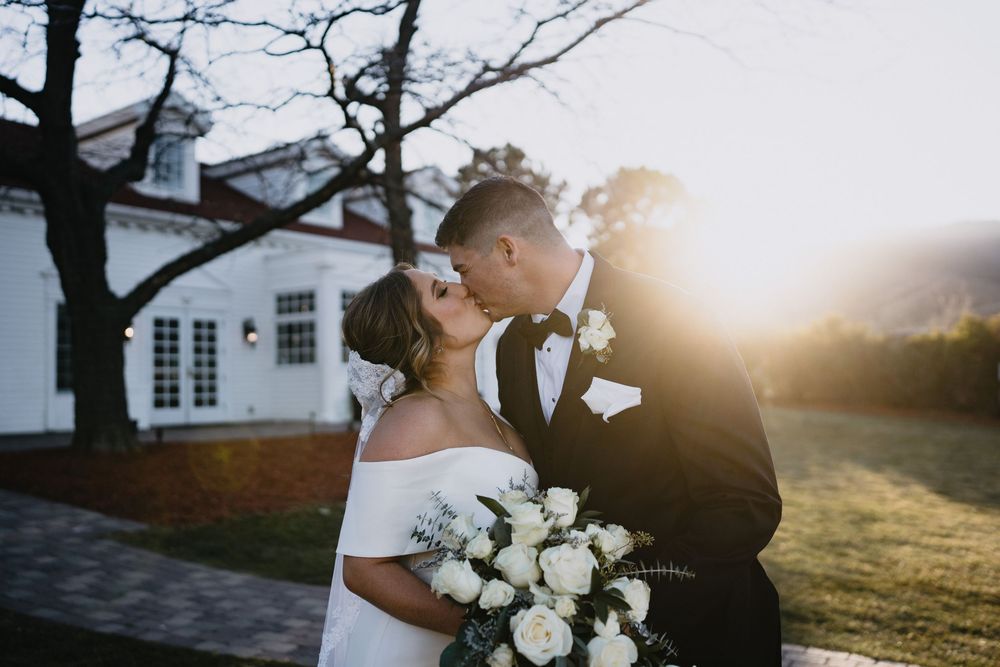 Bride and groom share a romantic kiss at golden hour, with bride holding white rose bouquet outside a white venue.