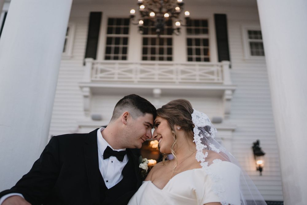 Bride and groom share a romantic moment, touching foreheads in front of a white Southern mansion with chandelier.