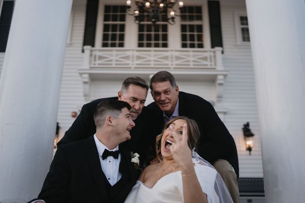 Joyful wedding couple laughing with two groomsmen on white mansion steps, bride in strapless gown, groom in black tuxedo.
