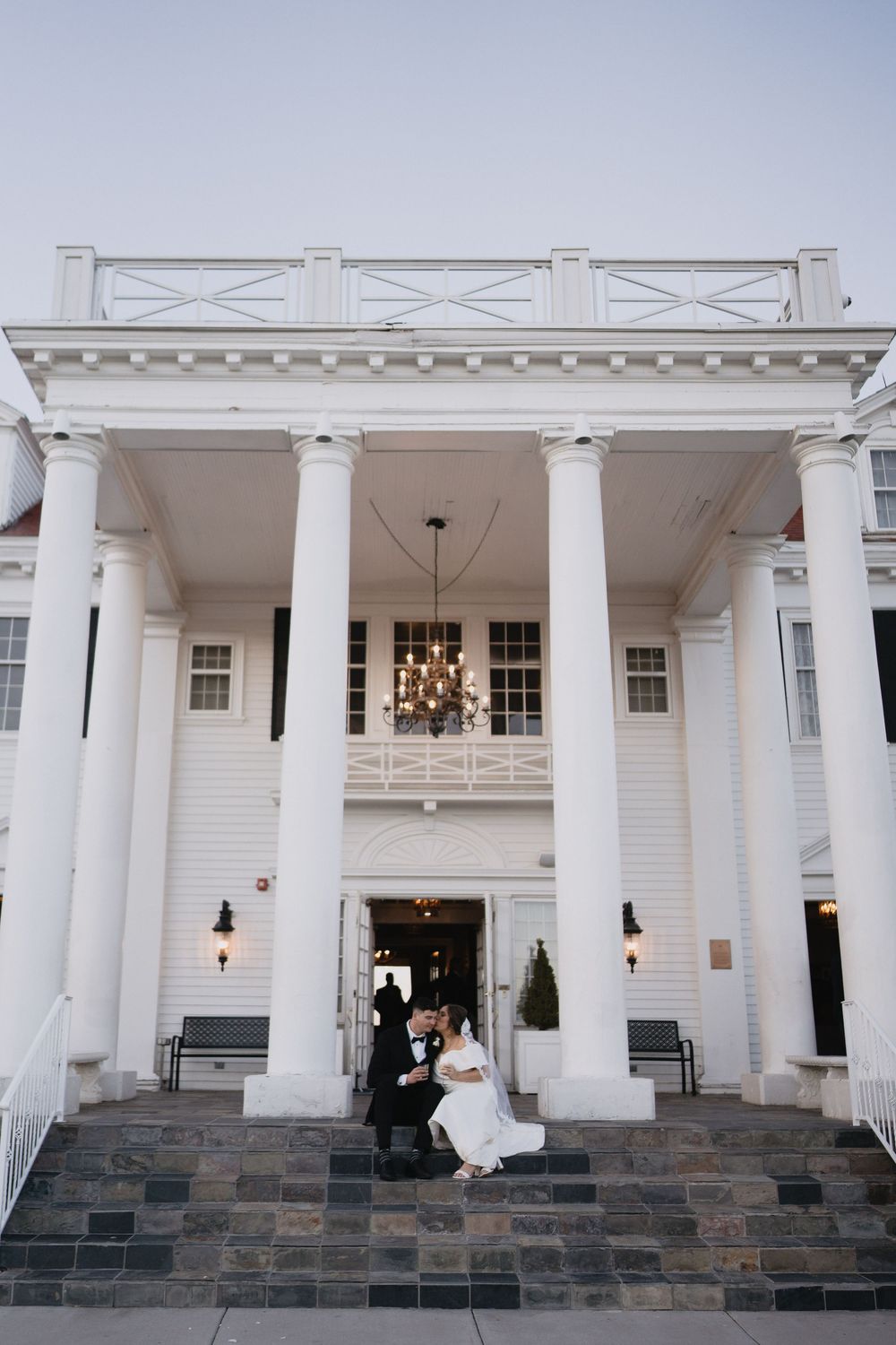 Bride and groom share a romantic kiss on the steps of an elegant white colonial wedding venue with grand columns.