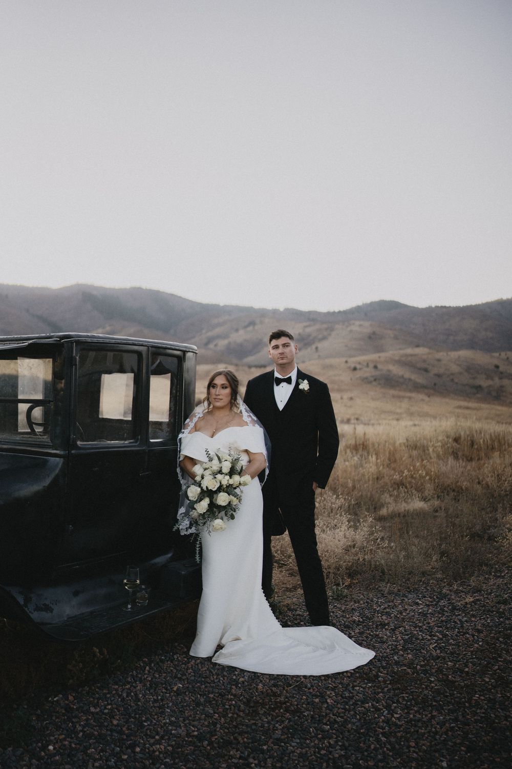 Bride and groom pose beside a vintage black truck with rolling hills in the background on their wedding day.