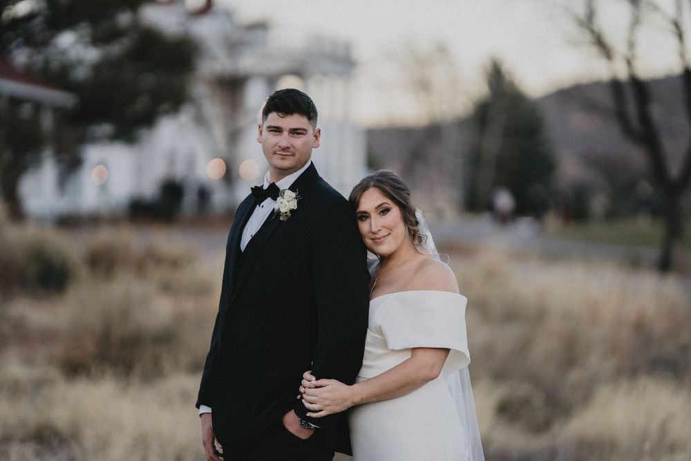Bride in off-shoulder white gown and groom in black tuxedo pose together outdoors at elegant wedding venue.