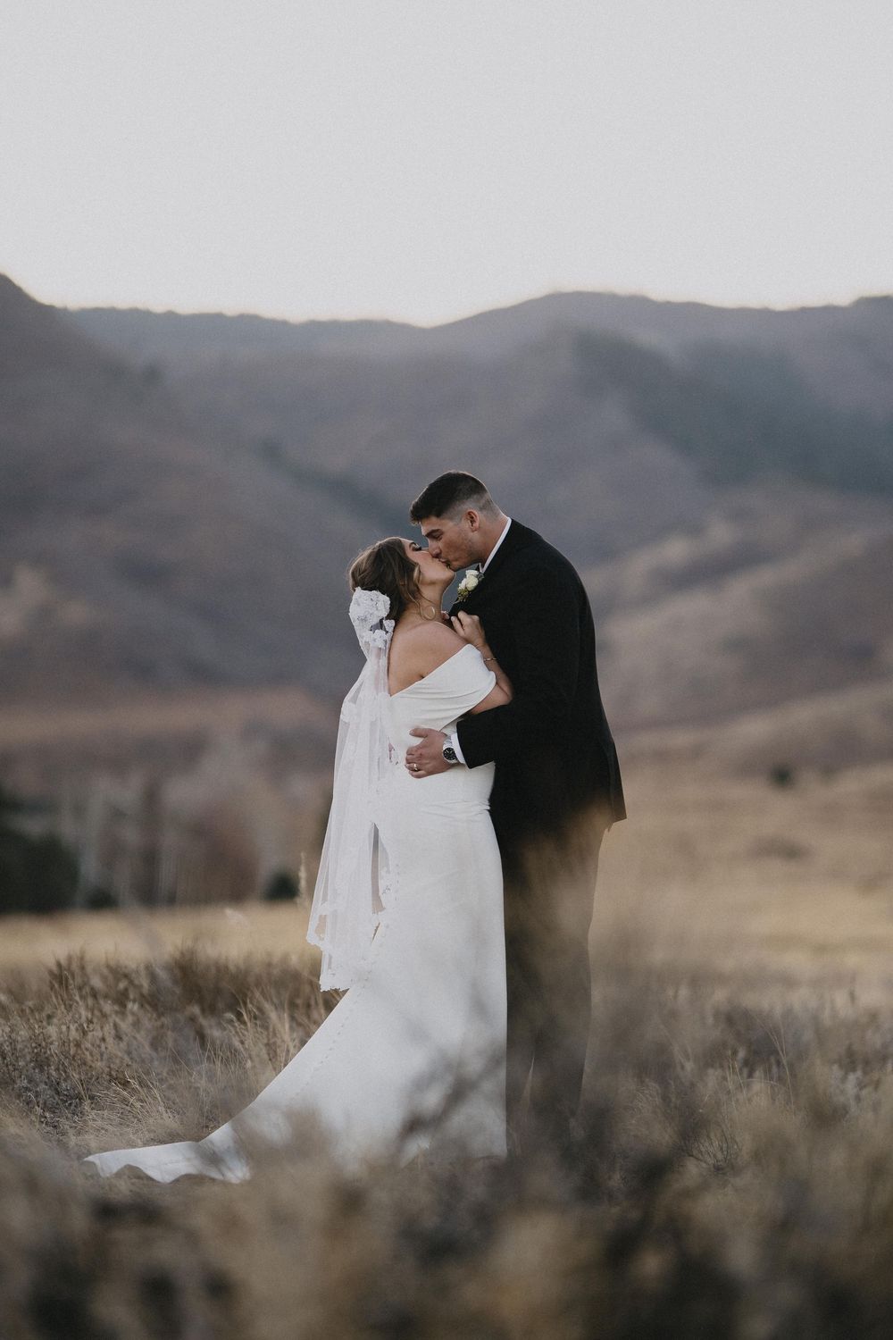 Bride and groom sharing a romantic kiss in a scenic mountain meadow, bride wearing an elegant white off-shoulder gown.