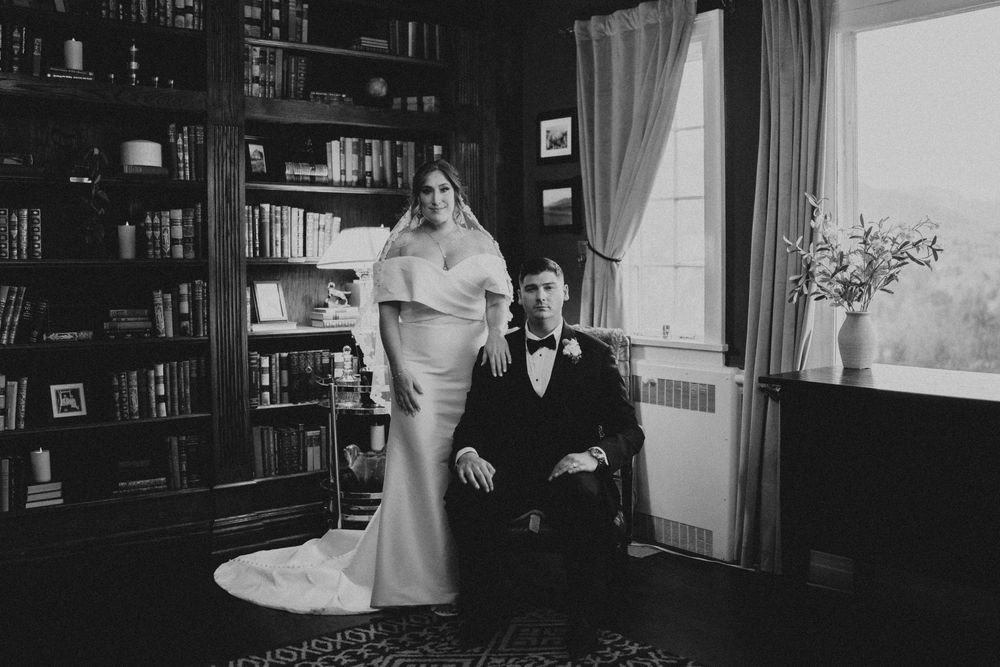 Black and white wedding photo of bride standing beside seated groom in elegant library with bookshelves.