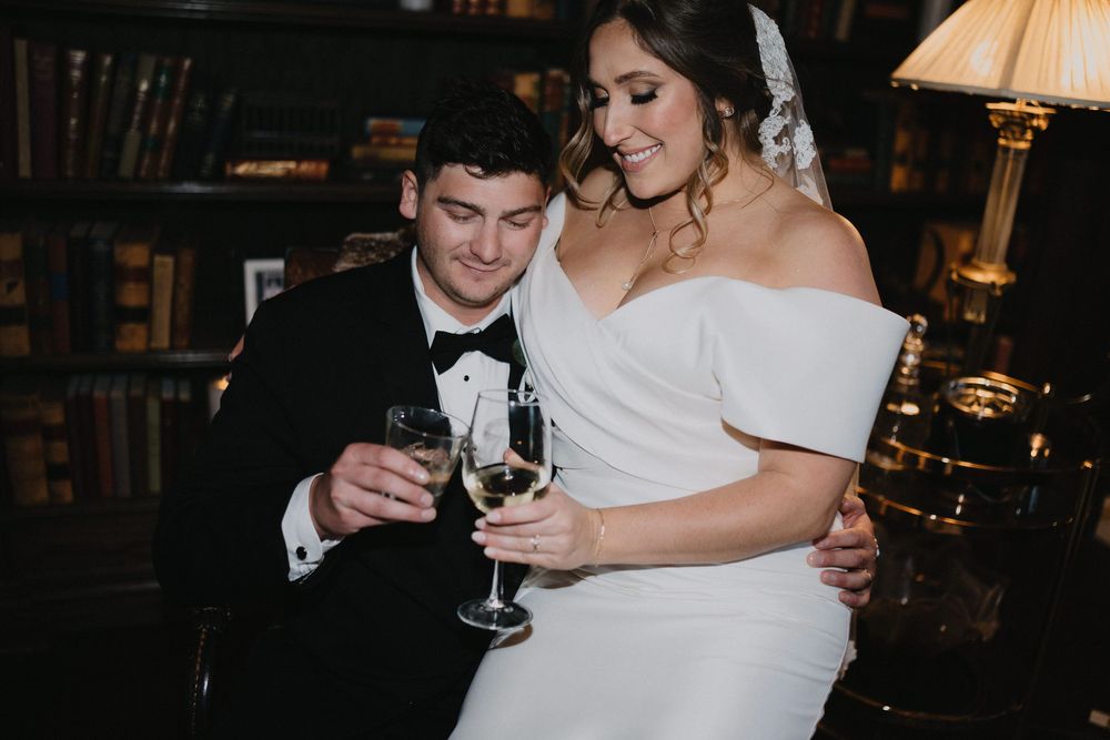 Bride and groom toasting with champagne glasses, bride in white off-shoulder gown, groom in classic black tuxedo.