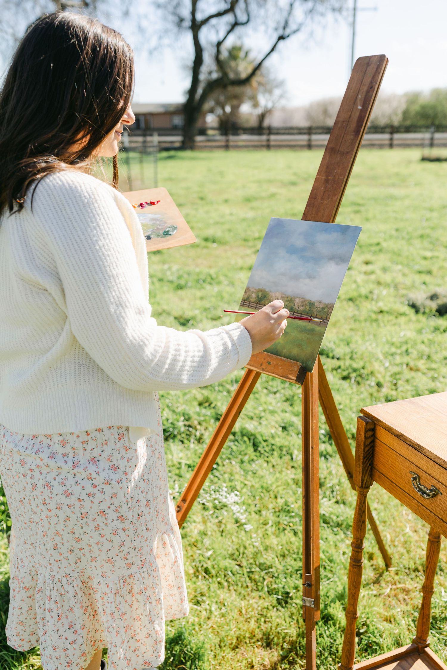 A young woman in a white sweater and floral skirt painting a landscape on an easel outdoors on a sunny day.