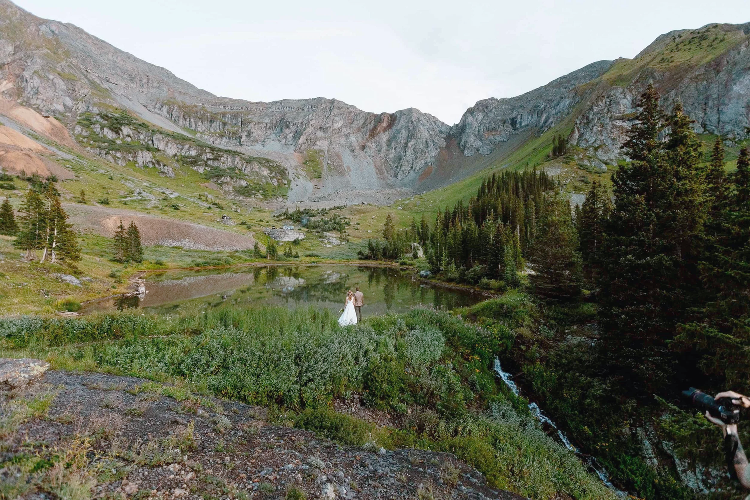 Groom lifting bride in white dress at mountain meadow wedding with pine trees and rocky peaks backdrop.