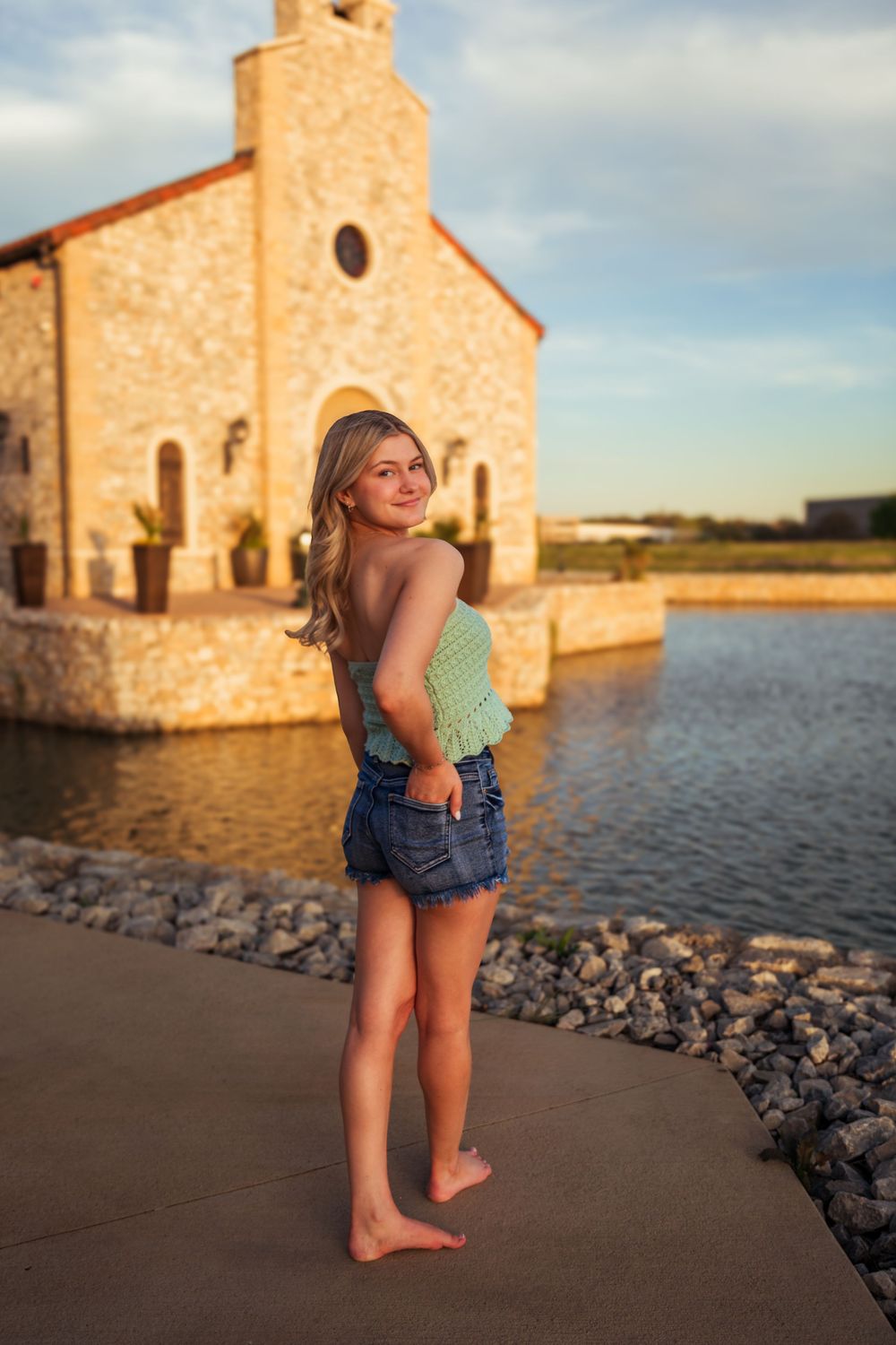Young blonde woman in green top and denim shorts poses barefoot by a stone chapel near a lake at golden hour.