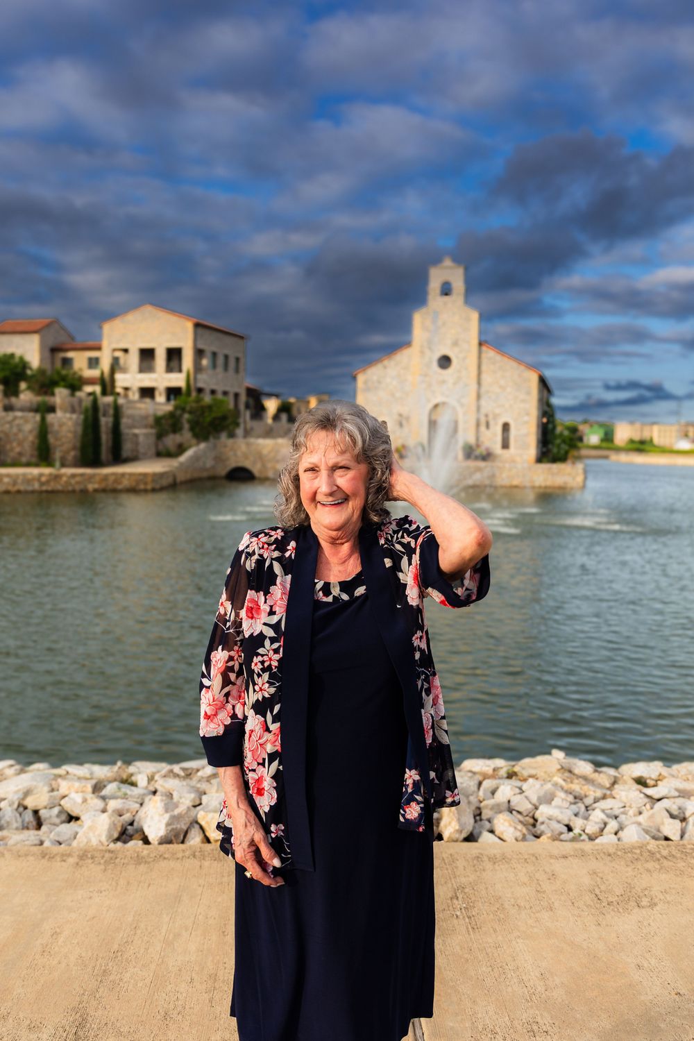 Smiling elderly woman in floral jacket poses by a scenic lake with a stone chapel fountain backdrop.