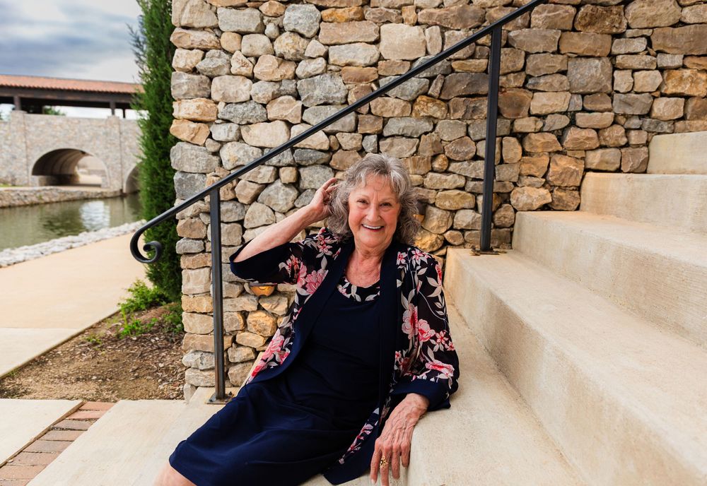 Elderly woman in floral jacket sits on stone steps outdoors near a rustic stone wall and bridge.