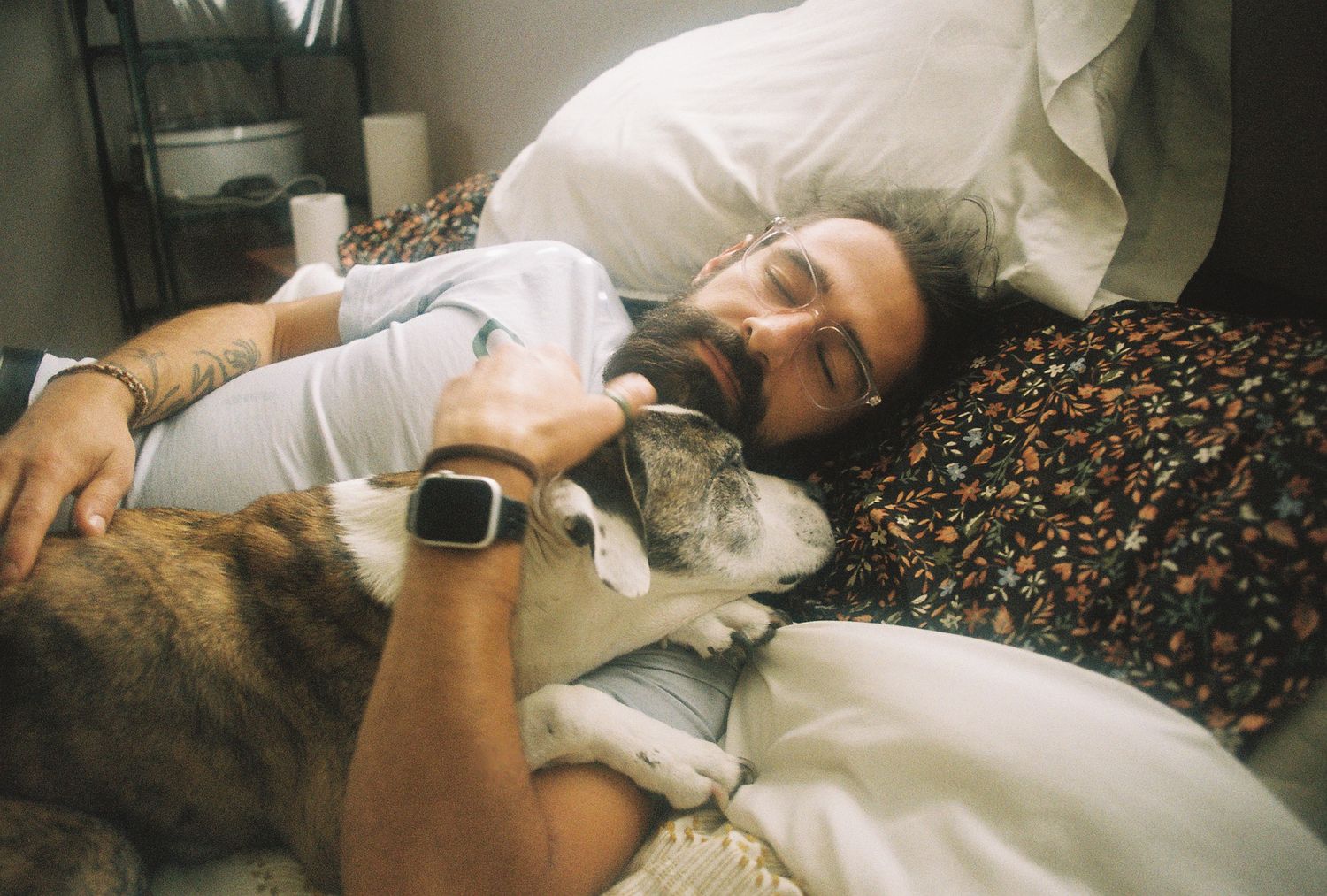 Bearded man cuddling with a dog in bed, wearing an Apple Watch in a cozy bedroom setting.