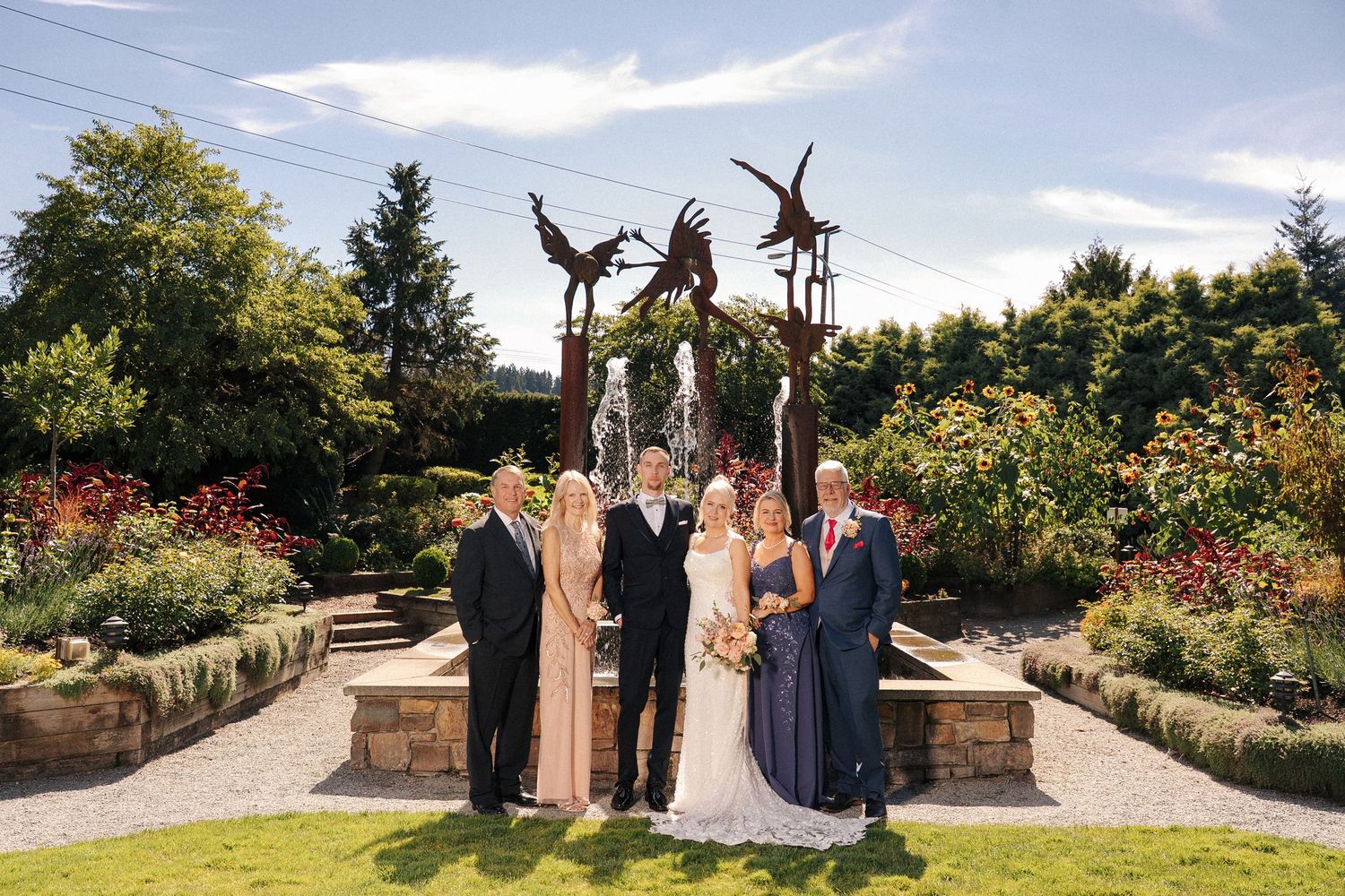 Wedding party poses before a bird sculpture fountain in a lush garden on a sunny day.
