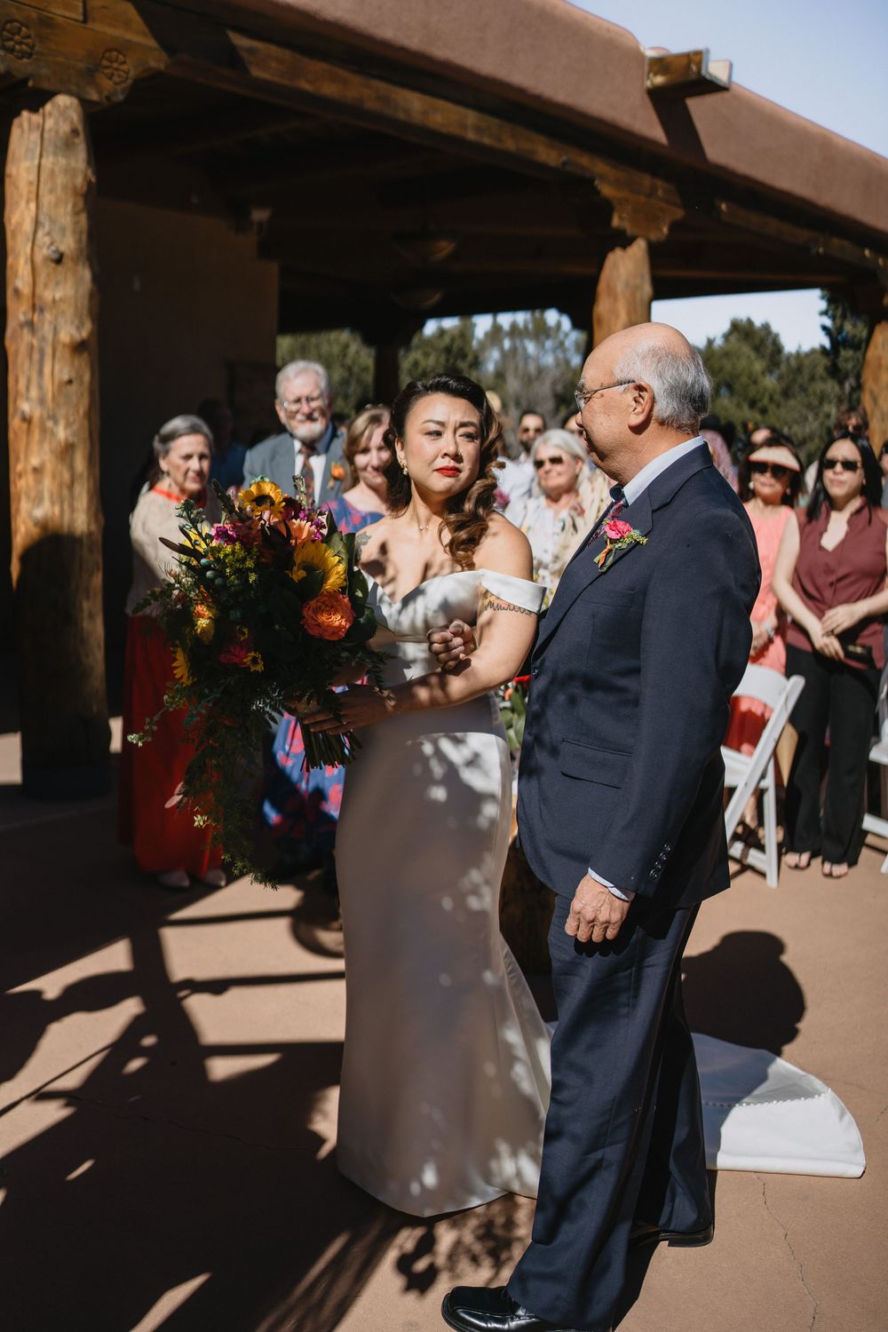 Bride in white off-shoulder gown holding vibrant bouquet walks down aisle with father.