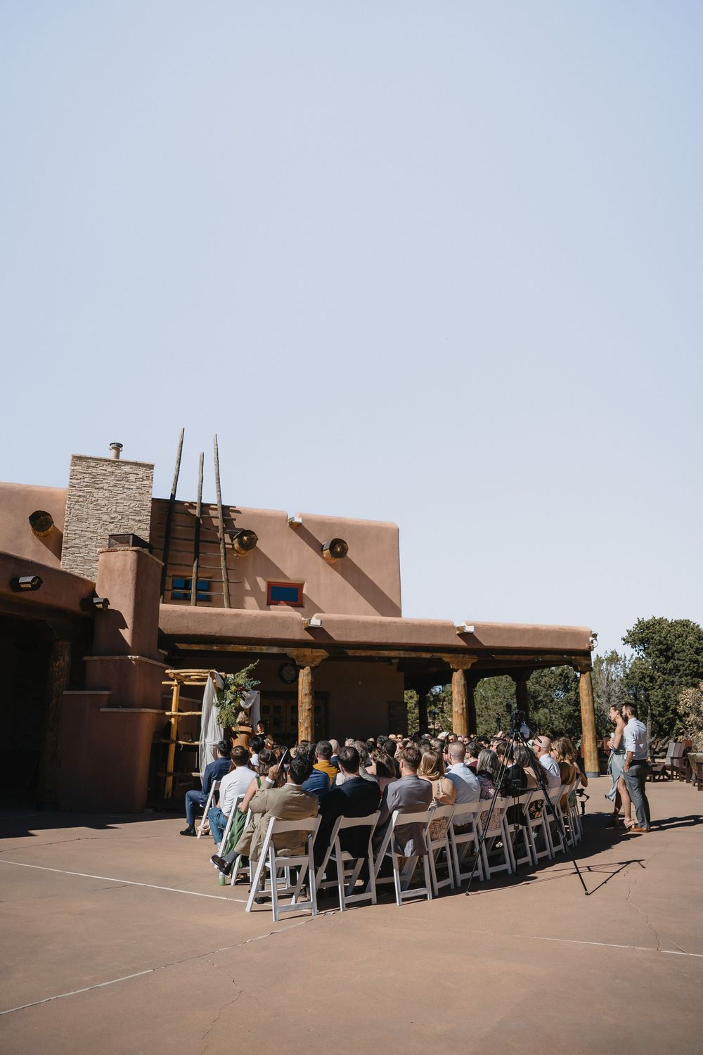 Guests seated at outdoor wedding ceremony beside rustic adobe Southwestern-style venue under blue sky.