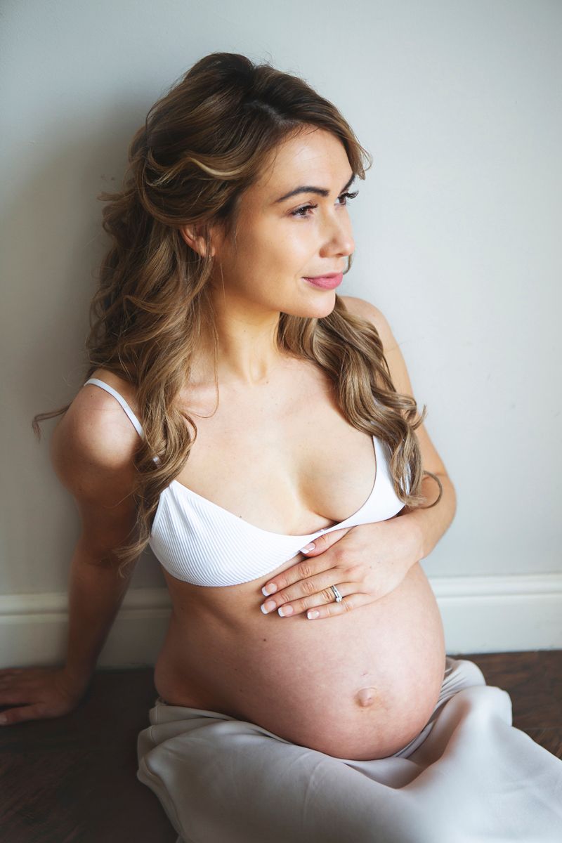 Pregnant woman in white bikini top cradling baby bump, sitting against wall during a at home maternity photoshoot in South East London