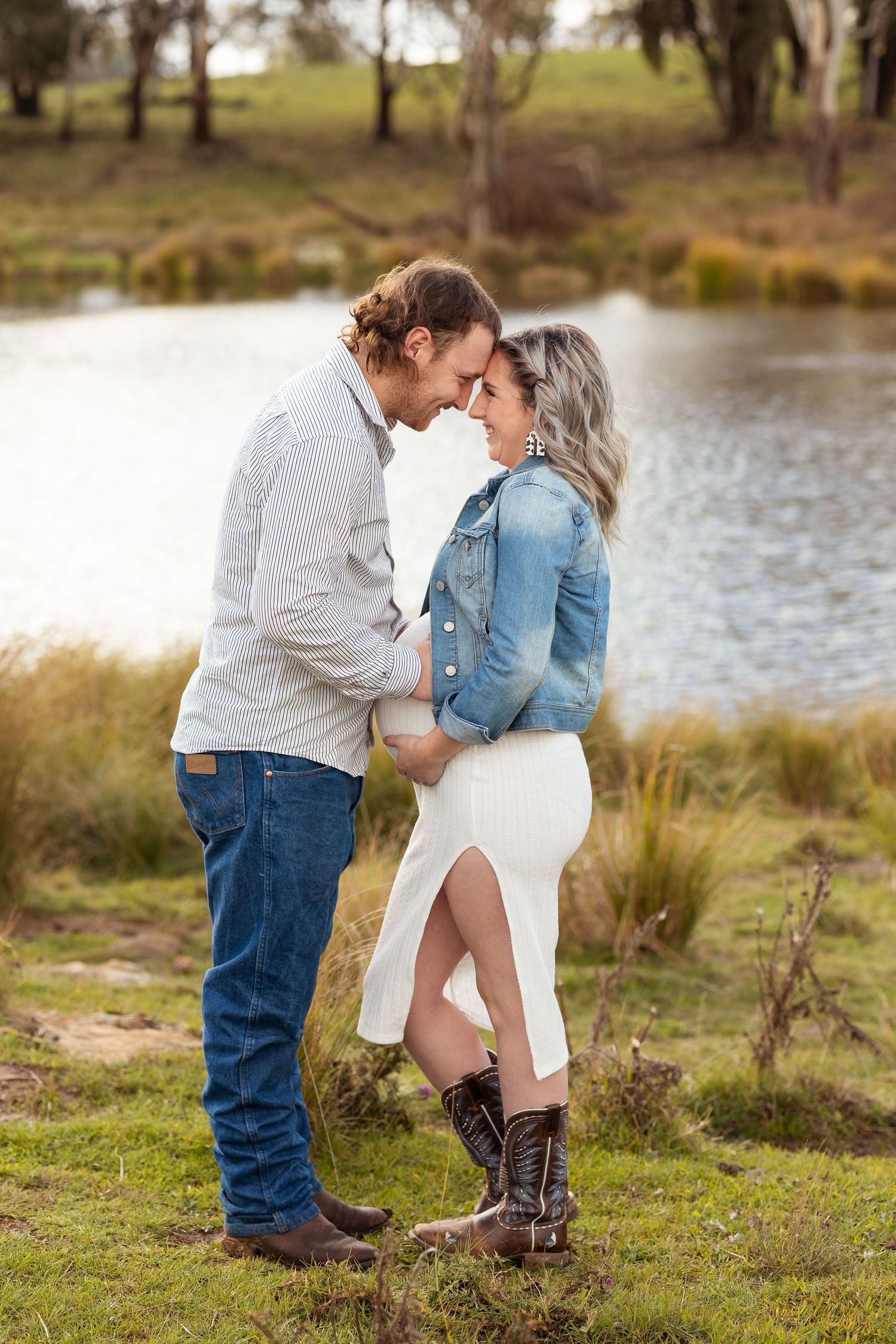 Romantic couple touching foreheads tenderly by a serene pond in casual country engagement photo.