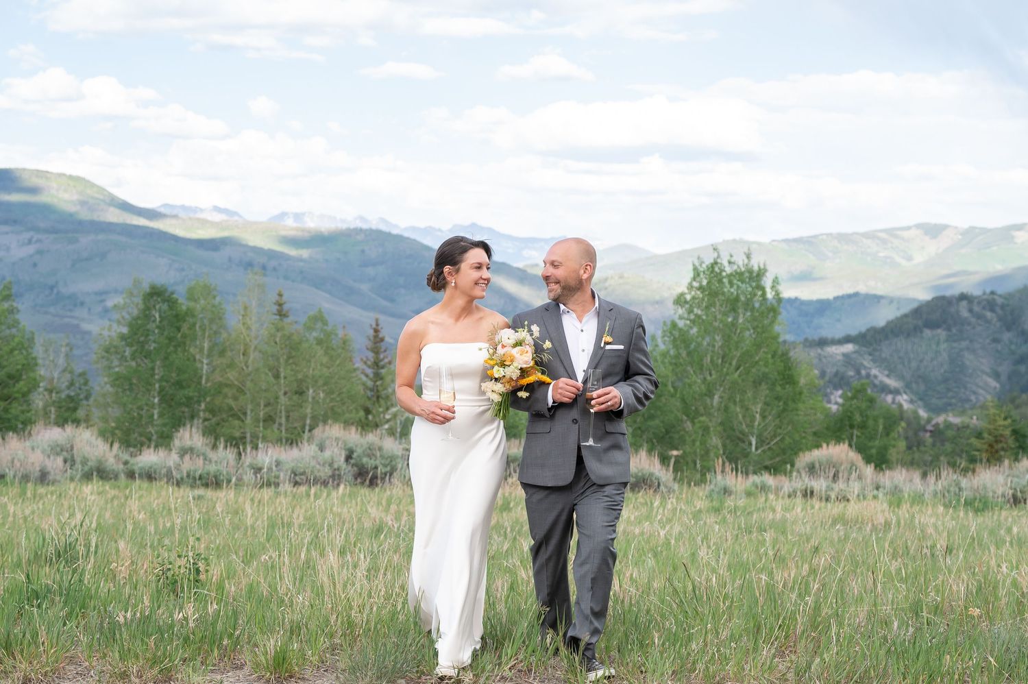 Bride and groom walking in mountain meadow, bride in white gown, groom in gray suit, scenic Colorado mountains backdrop.