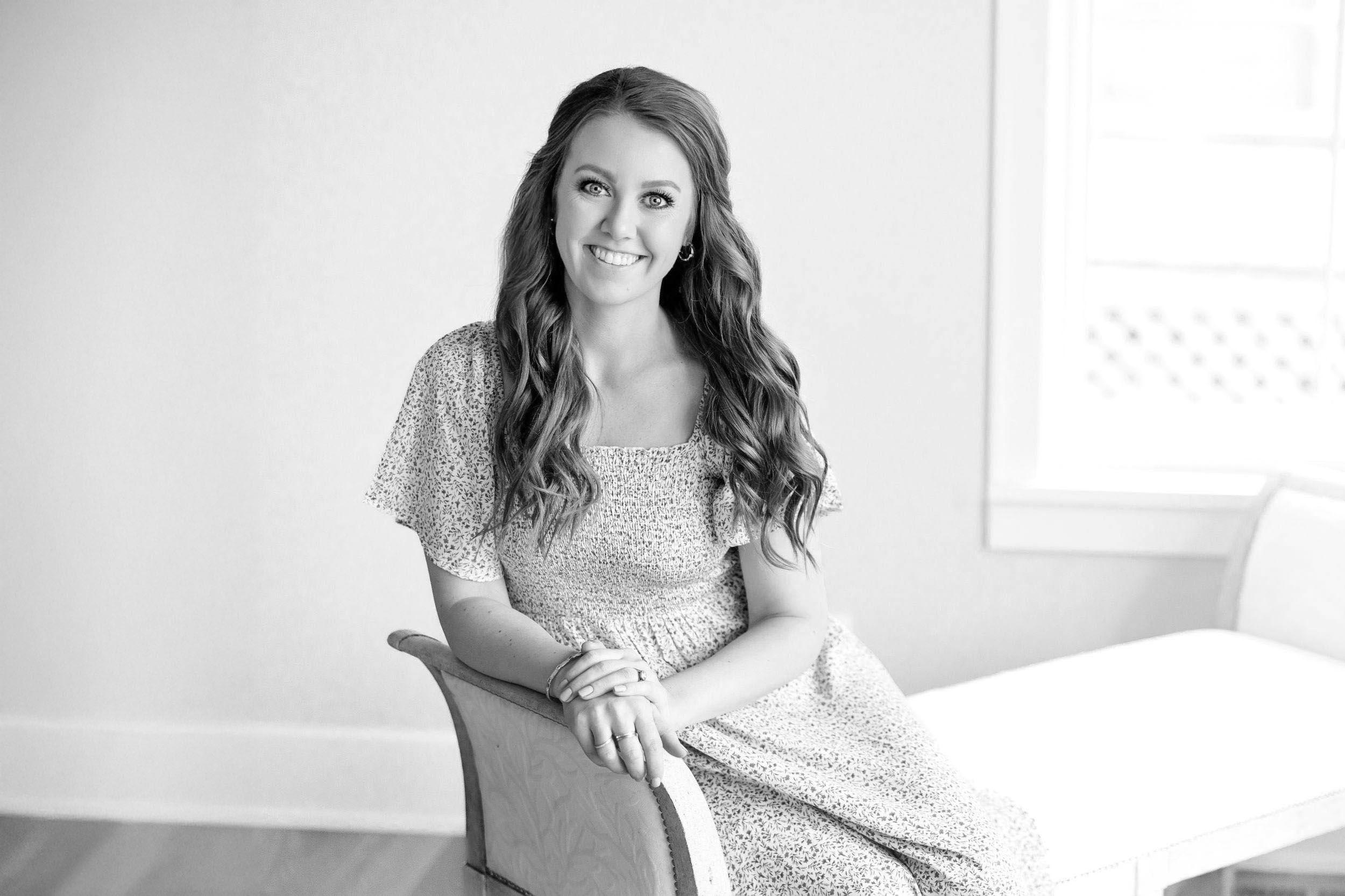 Black and white portrait of a smiling young woman in a floral dress seated gracefully in a chair.