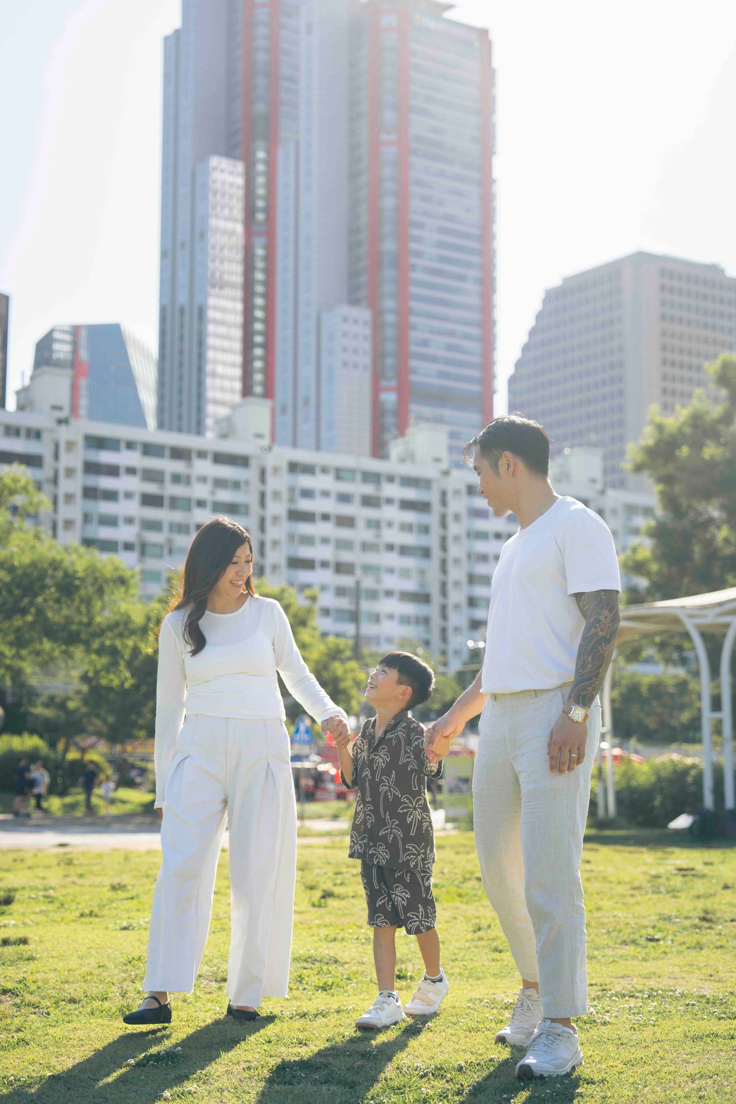 Asian family of three holding hands on green park lawn with city skyscrapers in sunny background.
