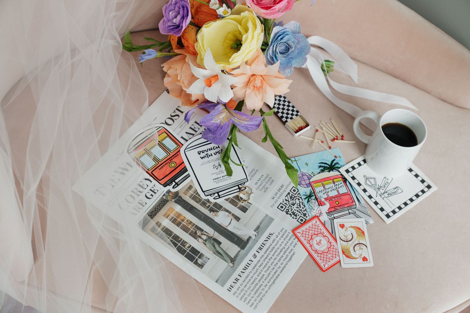 Colorful wedding flat lay with floral bouquet, coffee cup, magazine, playing cards, and ribbon on pink surface.