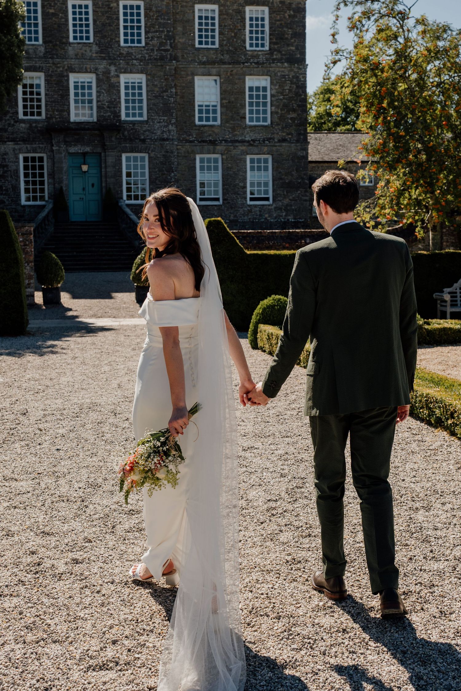 Bride in white gown holding bouquet walks hand-in-hand with groom in green suit on gravel path outside historic manor.