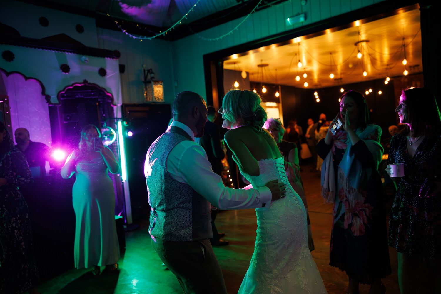 Bride and groom share first dance at wedding reception with green stage lighting and guests in background.