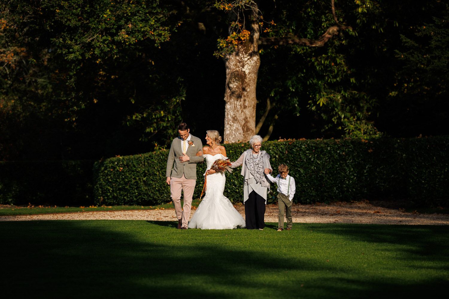 Bride in white gown walks with family on lush green lawn surrounded by trees at outdoor wedding.