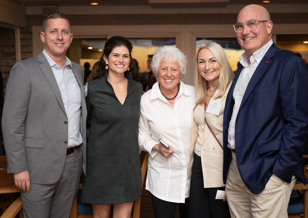 Five smiling adults posing together at a formal indoor event, dressed in business casual attire.