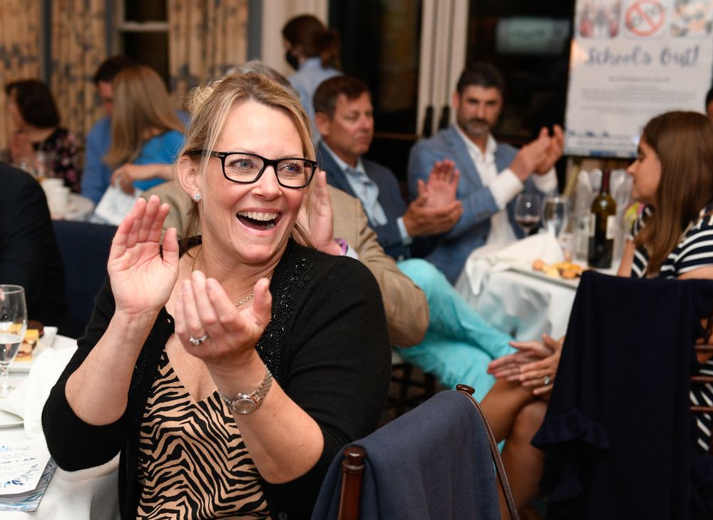 Woman in glasses laughing and clapping at a festive indoor event with guests applauding in the background.