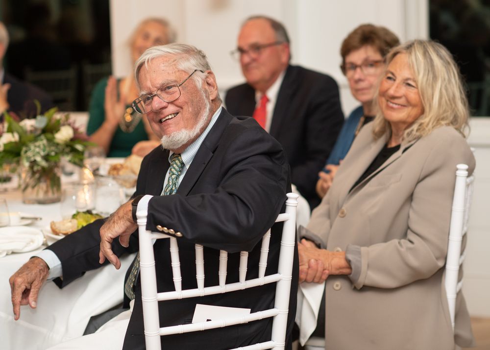 Elderly couple smiling and laughing together at a formal dinner event, seated at a white decorated table.