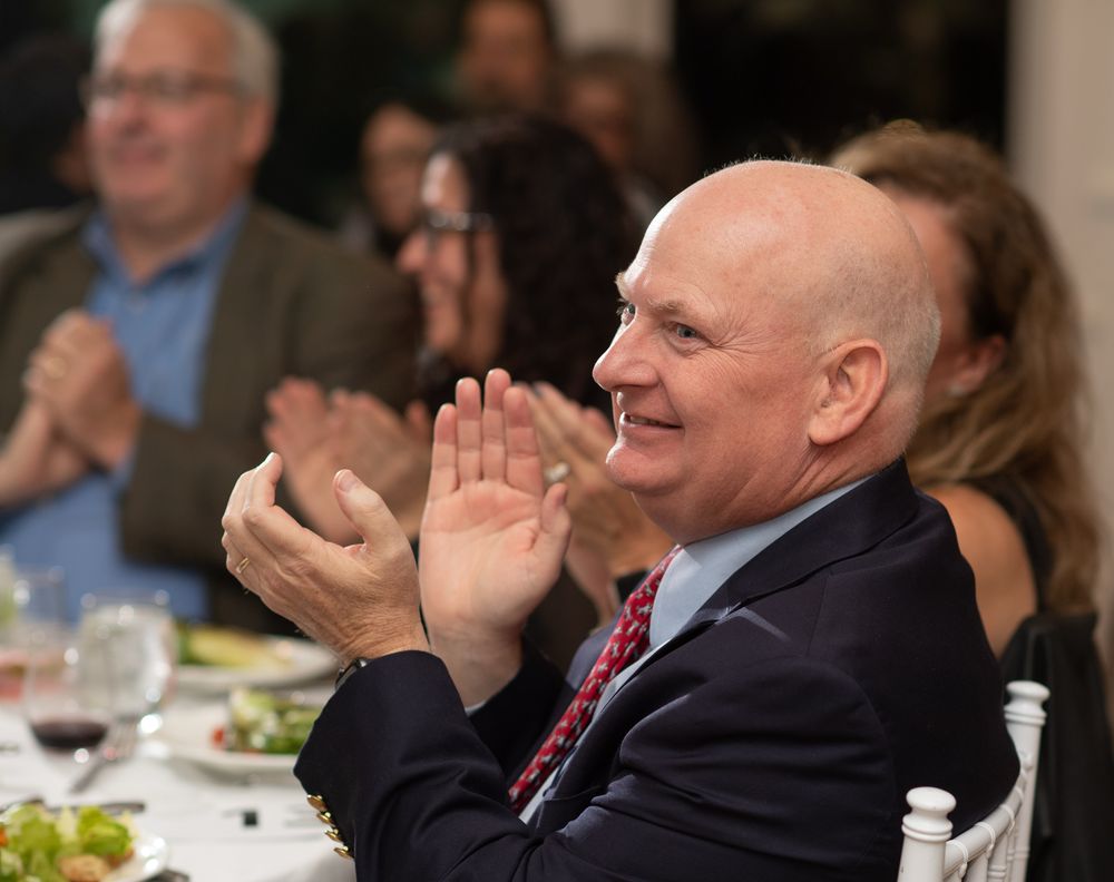 Smiling bald man in navy suit clapping at formal dinner event with guests applauding in background.