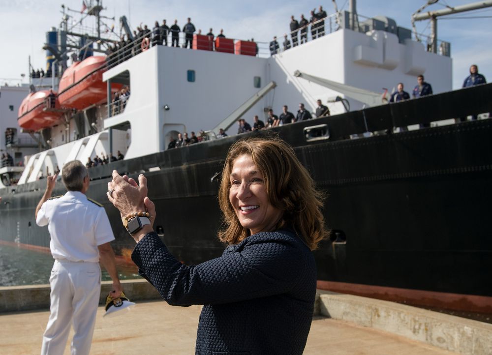 Woman smiling and taking photos near a large cargo ship docked at port with crew members visible on deck.
