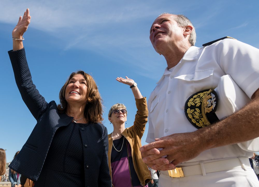 Two women and a naval officer in white uniform look upward outdoors on a sunny day.
