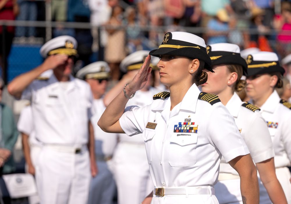 Female Navy officers in white uniforms saluting during a military ceremony outdoors.