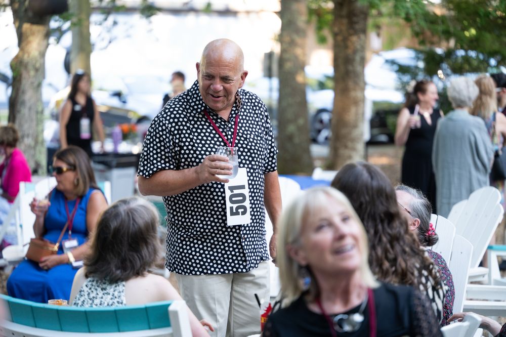 Bald man in polka dot shirt smiling and socializing at an outdoor event with guests seated nearby.