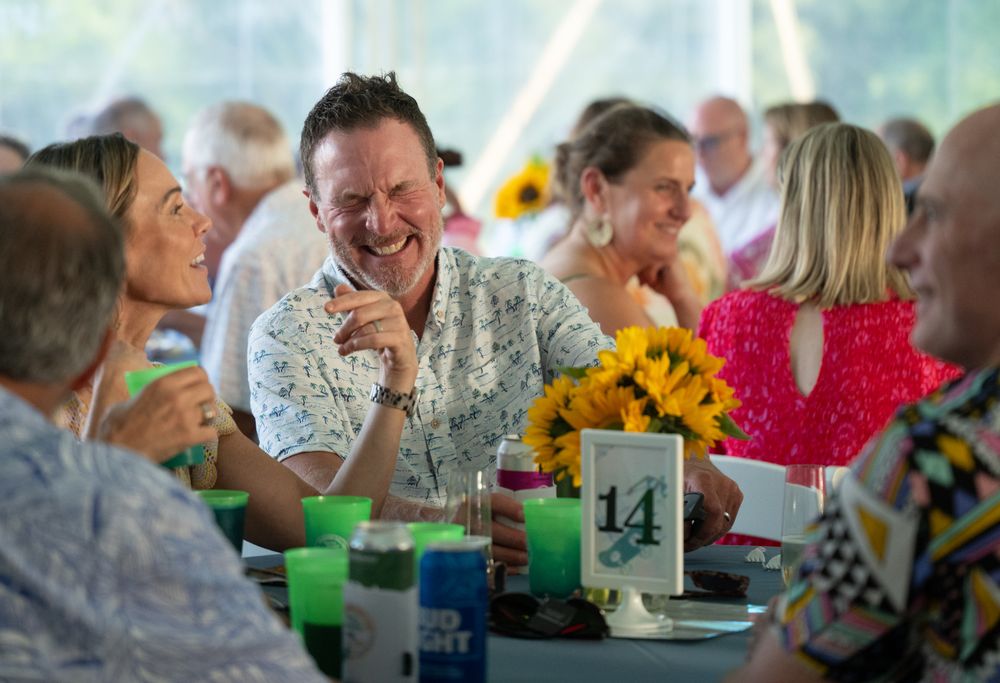 People laughing and socializing at an outdoor event under a tent with sunflowers and table decorations.