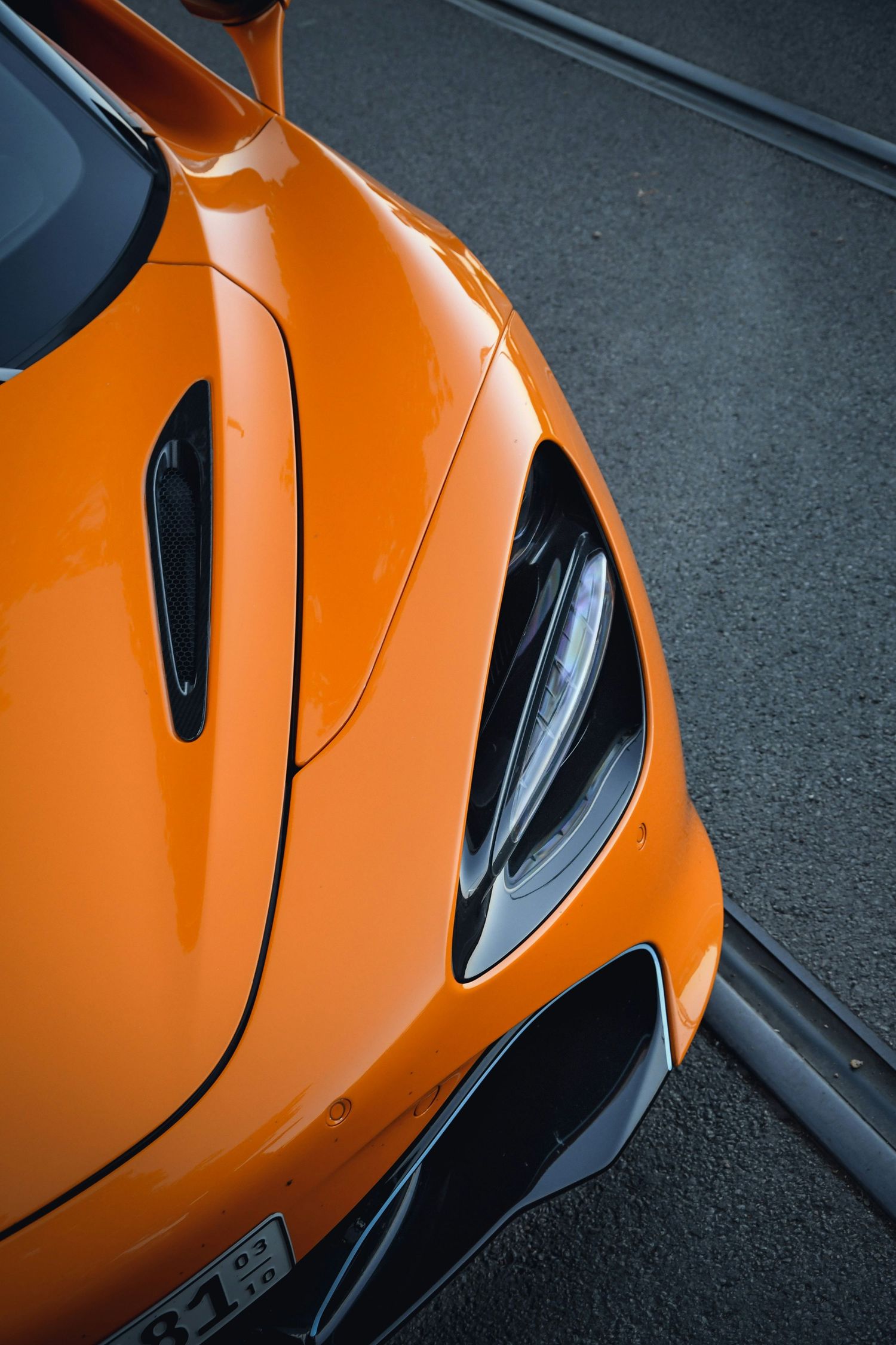 Close-up of an orange McLaren supercar front hood and headlight detail on a city street.